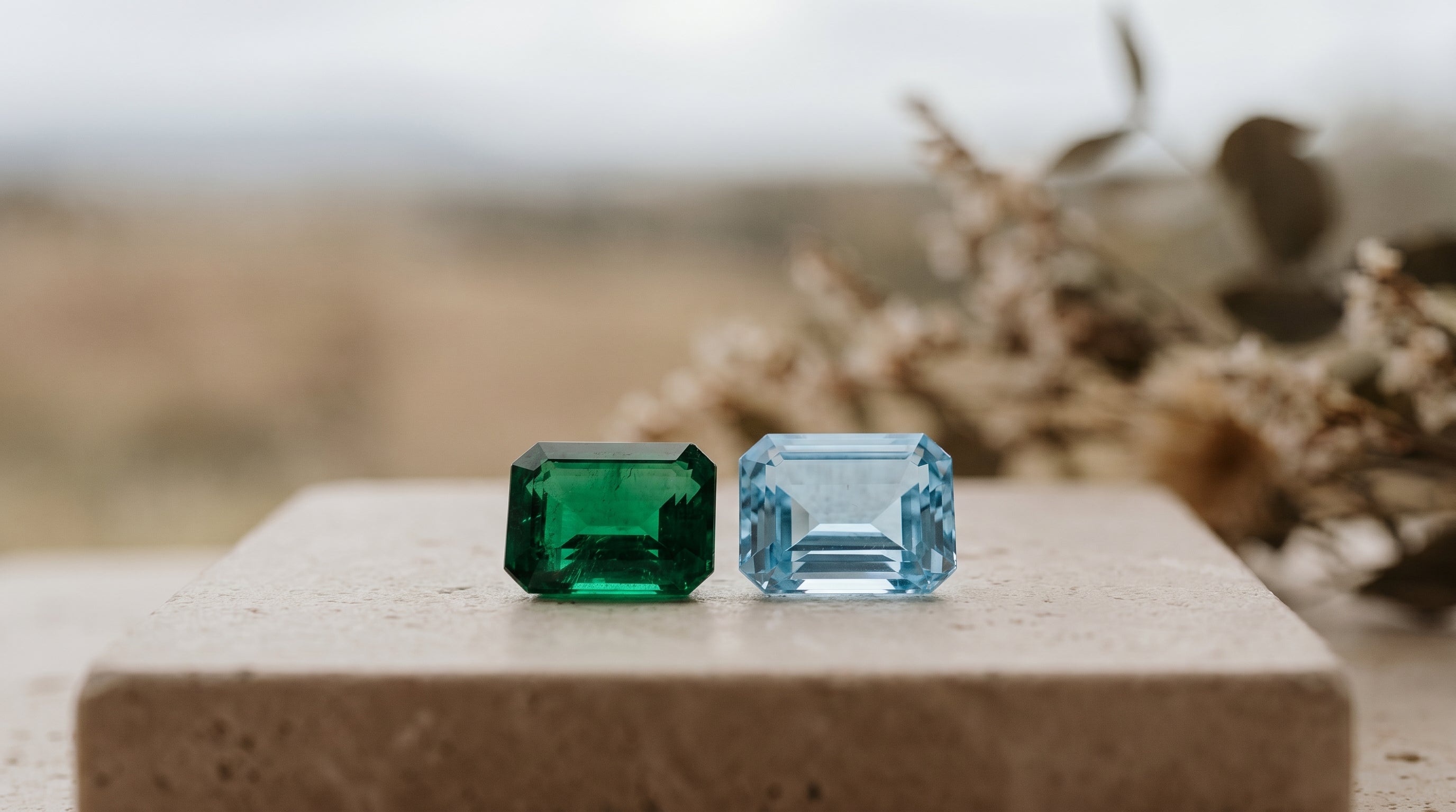 A faceted green emerald and a clear blue aquamarine side by side in soft natural light