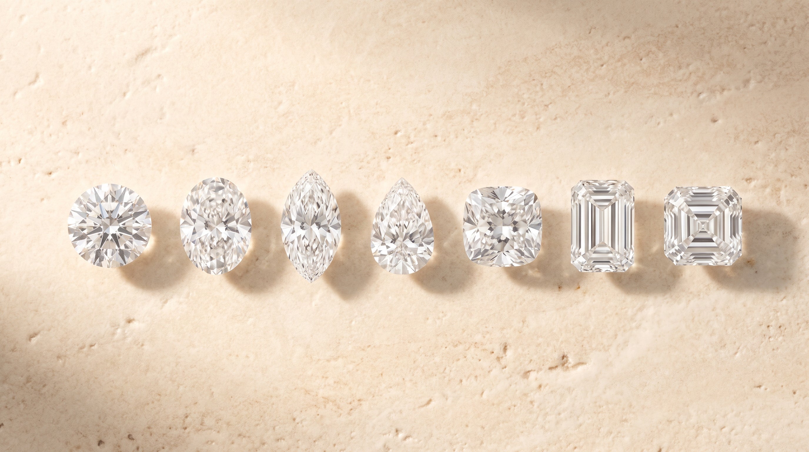 Seven loose diamonds in a row showing round, oval, marquise, pear, cushion, emerald and asscher cut shapes on travertine